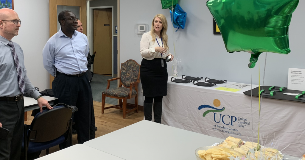 A woman speaking holding a device next to a table with equipment and an UCP banner. There are balloons and food on a table in the foreground. Three men stand listening. 