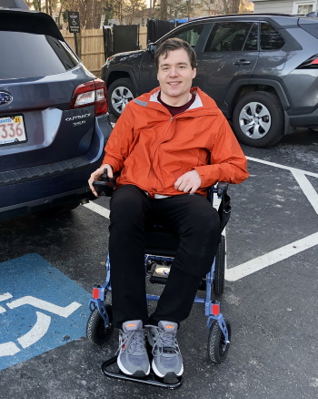 A man seated in a wheelchair smiling in a parking lot with accessible parking icon painted on pavement.